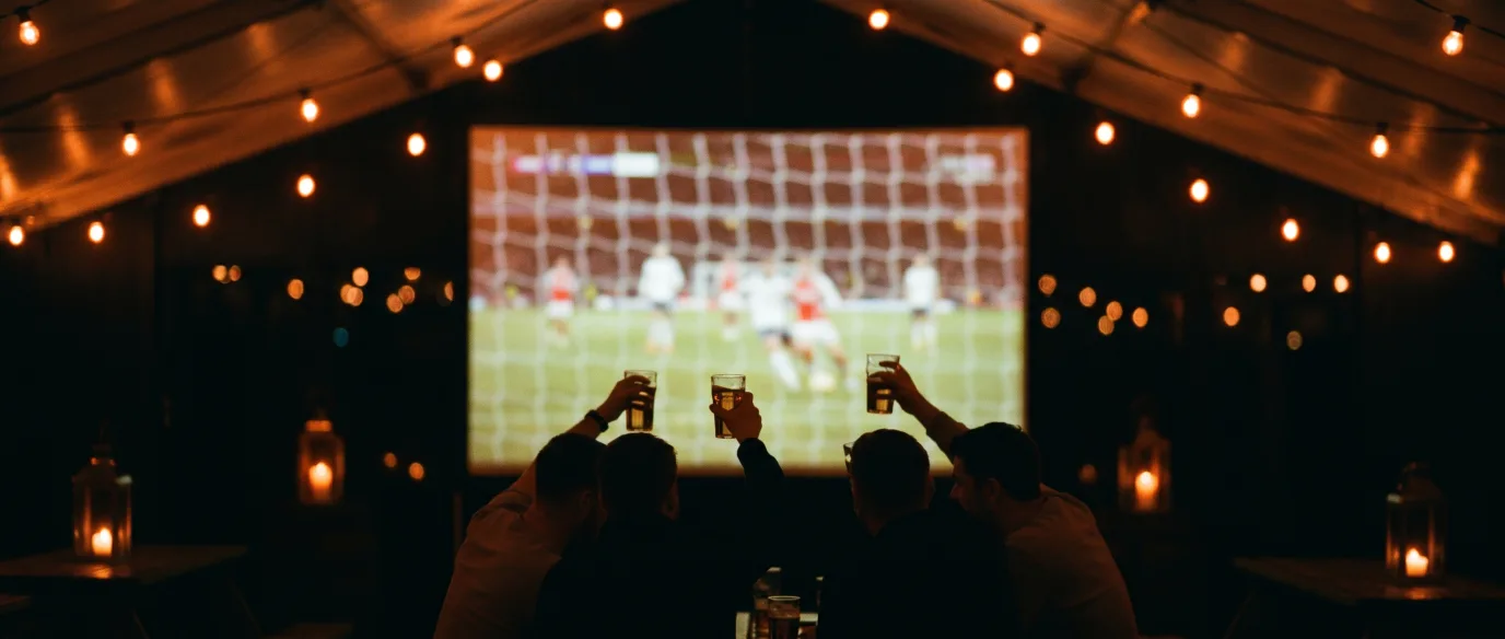 Football fans raising pints in the Fizzy Moon marquee in front of a big screen showing a match